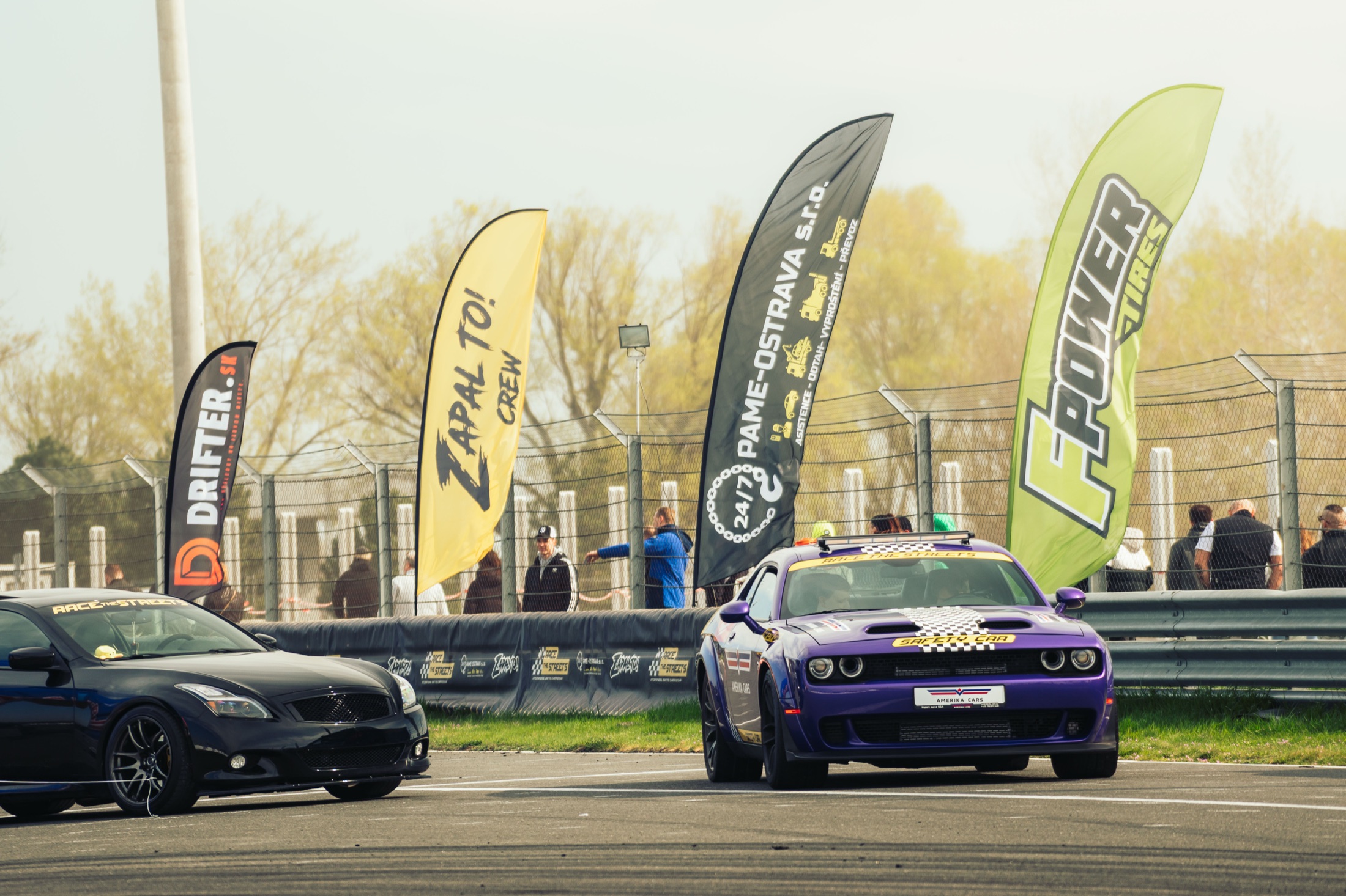 Žlutá plážová vlajka Zápal to! Crew mezi dalšími vlajkami u okruhu, na trati projíždí safety car